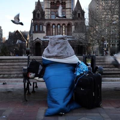 Bundled in blankets and a sleeping bag, a homeless person sits on a bench in Copley Square in Boston. 
