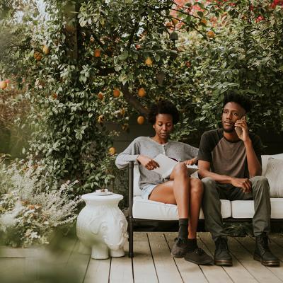 A black couple sitting on a bench in their home garden while reading a book and using a phone.
