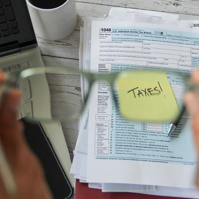 A selective focus of person holding eyeglasses over a tax form and the word "taxes" on a note.