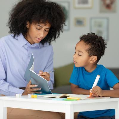 Black adult helping child learn and write, looking at a book.