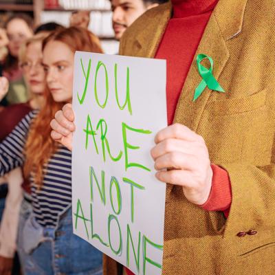 Young adults and a mental health counselor wearing a green ribbon and holding up a "you are not alone" sign to drive support during Mental Health Awareness month.