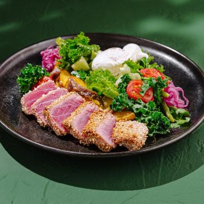 Seared tuna, coated in sesame seeds, is served on a black plate with fresh greens, colorful vegetables, and poached eggs, highlighting vibrant flavors and textures.