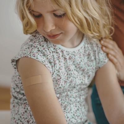 A young child with a small adhesive bandage on their upper arm. The child is wearing a floral-patterned shirt and looking down at the bandage.
