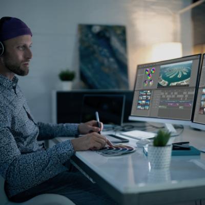 A man wearing a hat and headphones sitting in front of two large computer screens while holding a pen in one hand and pointing to the a color palette with his right index finger.