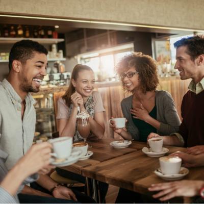 A group of six friends gathered around a wooden table in a cozy café. They are all smiling and laughing. Each person is holding a cup of coffee.