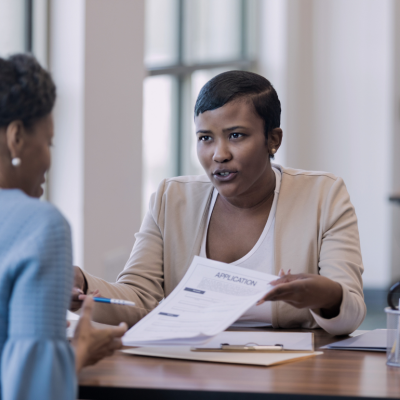A Black woman at the desk is showing an application document to an African American woman sitting opposite her.