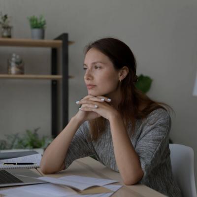 A woman sitting at a desk resting her chin on her hands with her fingers interlocked. She's looking forward. 