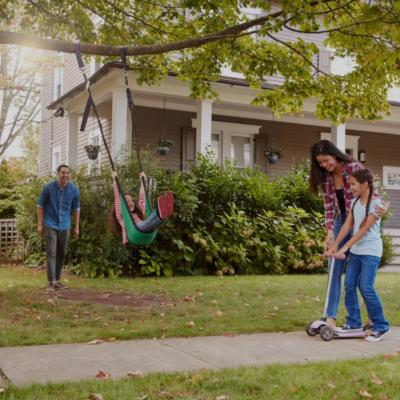 A family in front of a suburban home with a well-maintained lawn and a tree with a swing. A young girl is swinging while a man stands nearby with a smile, seemingly pushing. To the right, a woman is helping another young girl ride a scooter on the sidewalk. 