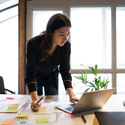 A business person stands at her desk and takes notes while reading her laptop.