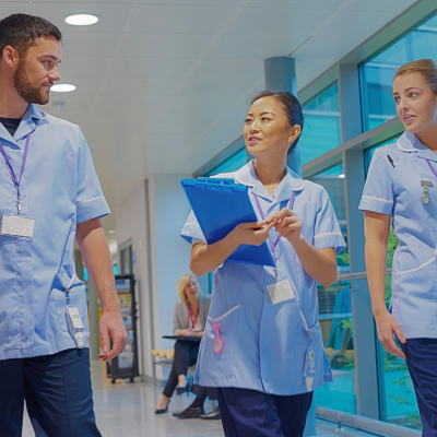 A male nurse and two female nurses walking in the hospital looking at the nurse in the center with a clipboard. 