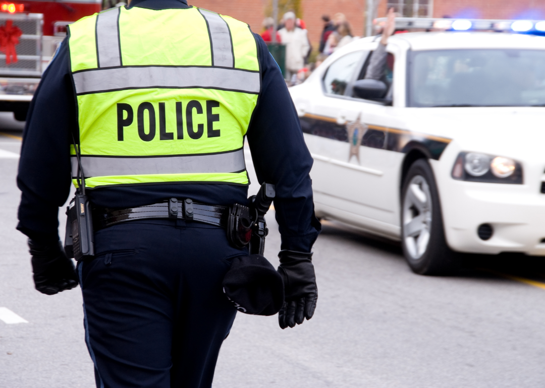 A police officer stands with his back to the camera, a white police car in the distance.