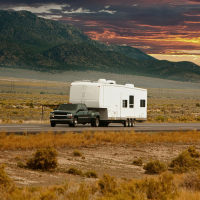 A black pickup truck towing a white camper trailer down a long highway.