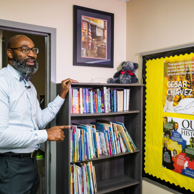 A teacher speaking in his classroom, pointing to posters featuring Black history icons figures and Cesar Chavez. 