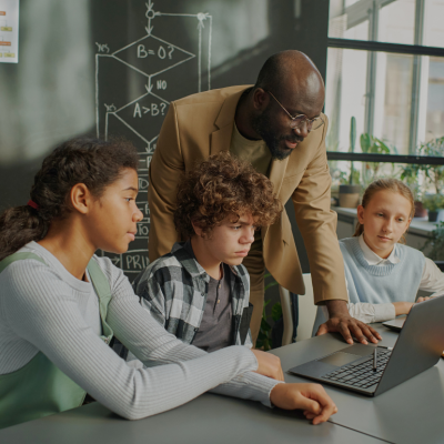A young Black male teacher looking at the computer screen on the desk, with two girls and a boy focused on the same screen.
