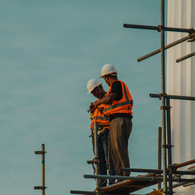 Two men in bright-orange vests wearing hard hats are standing on scaffoldings holding tools.