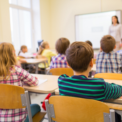A rear view of young students sitting and listening to teacher in a classroom