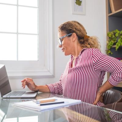 A woman sitting at a desk, grimacing and holding her hand on her back.