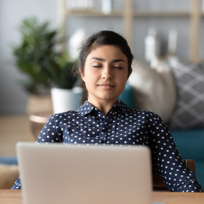 A calm-looking woman sitting with her eyes gently closed in front of a laptop in a home office.