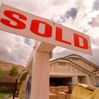 Close-up of a sign reading "Sold" in bright orange in front of a house and lawn full of boxes.