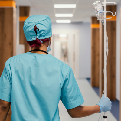 A worker in green scrubs walks down a hospital hallway pushing an IV bag.