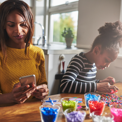 A woman checking her phone while a little girl does crafts next to her.