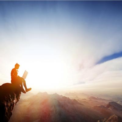 Man sitting on a cliff working on a laptop.