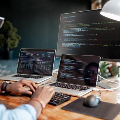 Young male programmer writing code with three monitors on table.