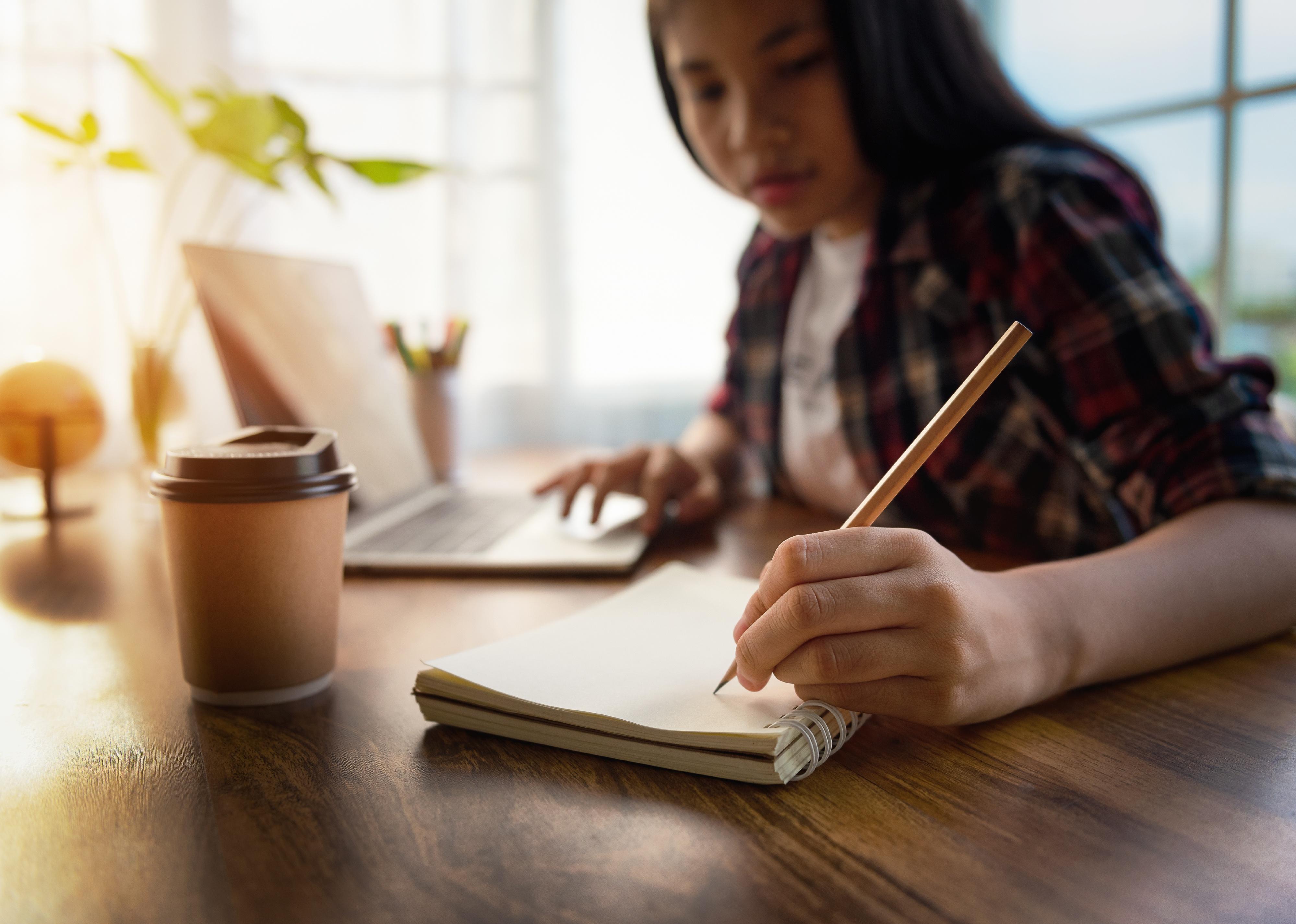 Student writing in a notebook and using laptop for homework.
