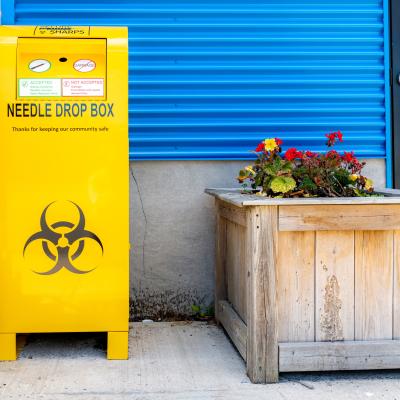 A public drop box for used needles, on a sidewalk by a building.