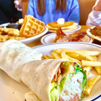 A table at IHOP with plates of brunch items, including a chicken and bacon wrap with fries, chicken and waffles, bacon, and a stack of pancakes.