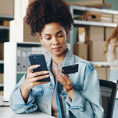 Black woman entrepreneur looking at a business credit card and holding smartphone inside an office with boxes and shelves and a blurred blond person in the background. 