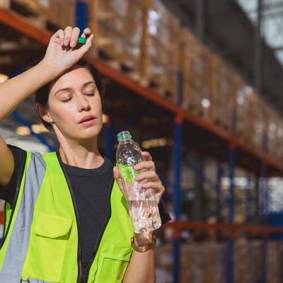 Woman wearing a high visibility vest holds a bottle of water and brushes off her forehead while her eyes are closed; stacks of pallets in a warehouse in the background.