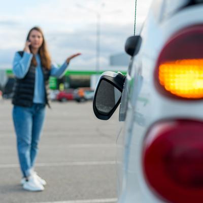 Woman talking on her phone while looking at her car's broken left side rear-view mirror.