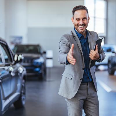A friendly and cheerful salesman poses to greet car showroom clients with a handshake.