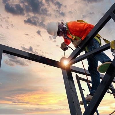 Low angle view of a welder is welding steel on a steel roof truss with the sunrise in the background.