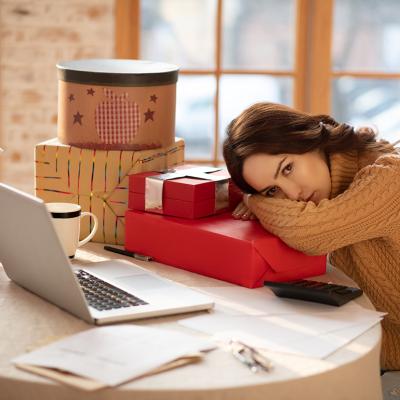 A young woman resting her head on a Christmas gift box alongside other presents in front of her laptop.