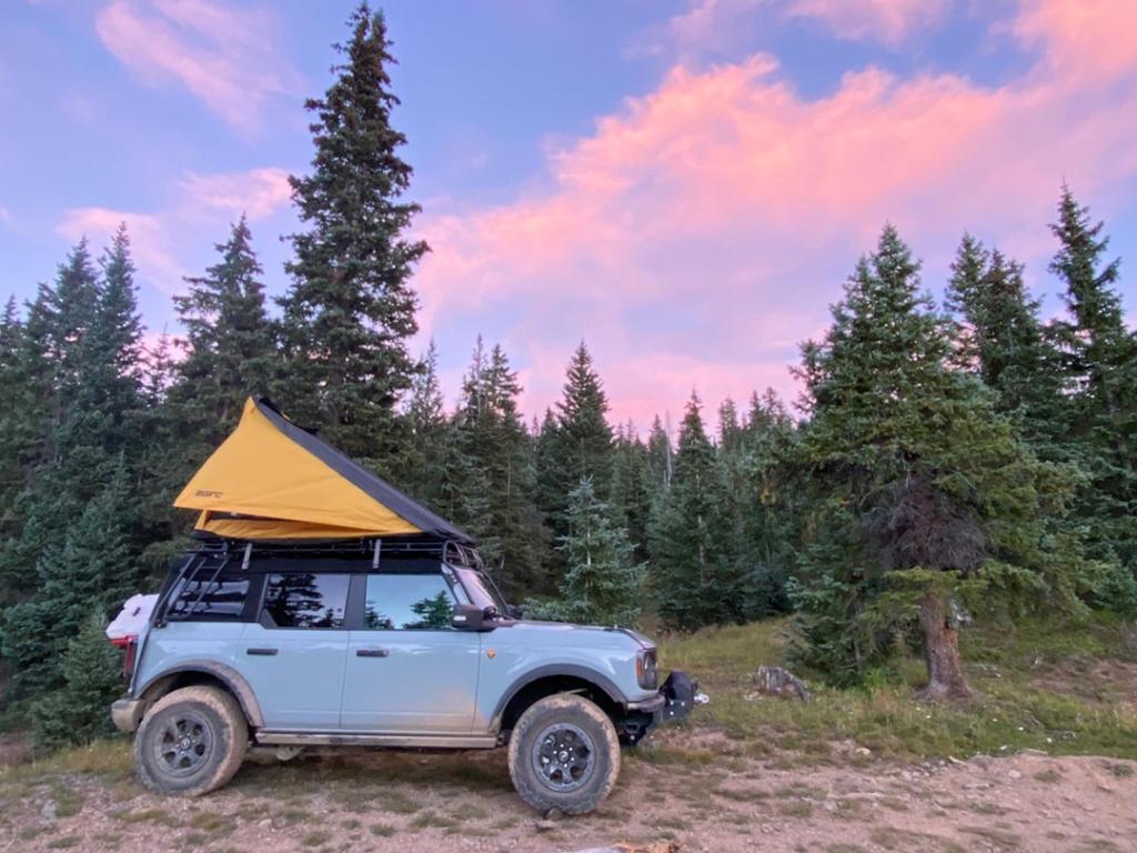 2021 Bronco blue Ford Bronco with canopy parked in dirt with pink clouds and blue sky and forest in background.