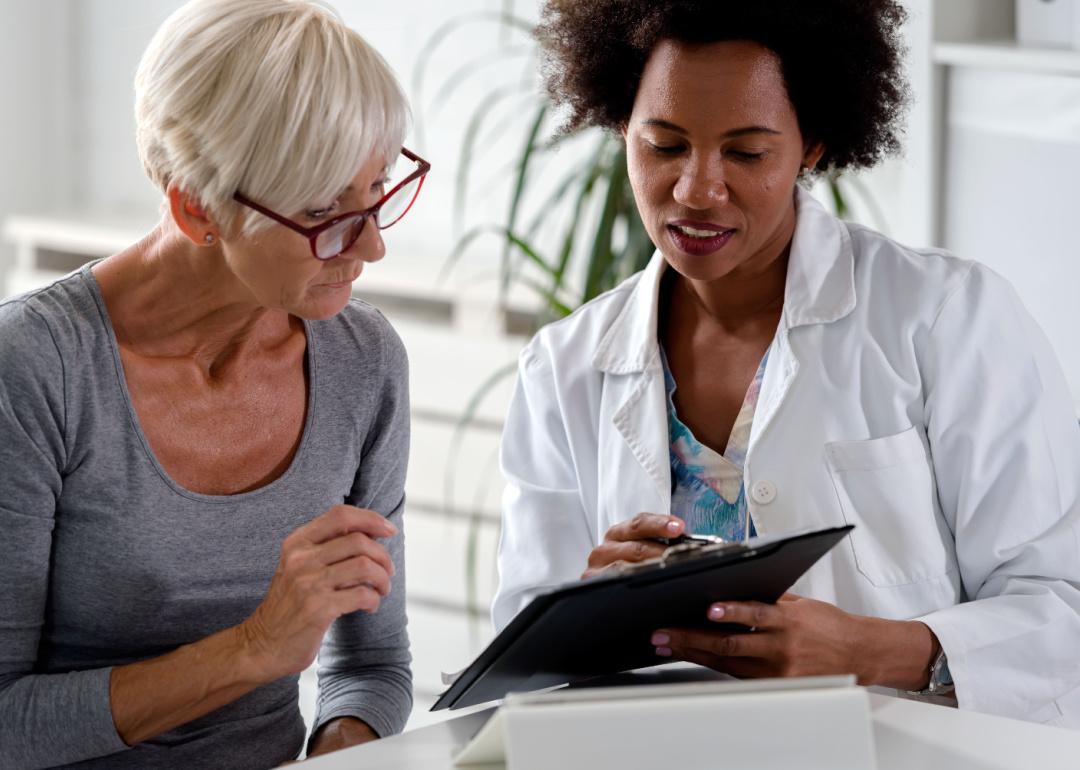 An older patient sits beside a doctor who is reviewing information on a clip board.