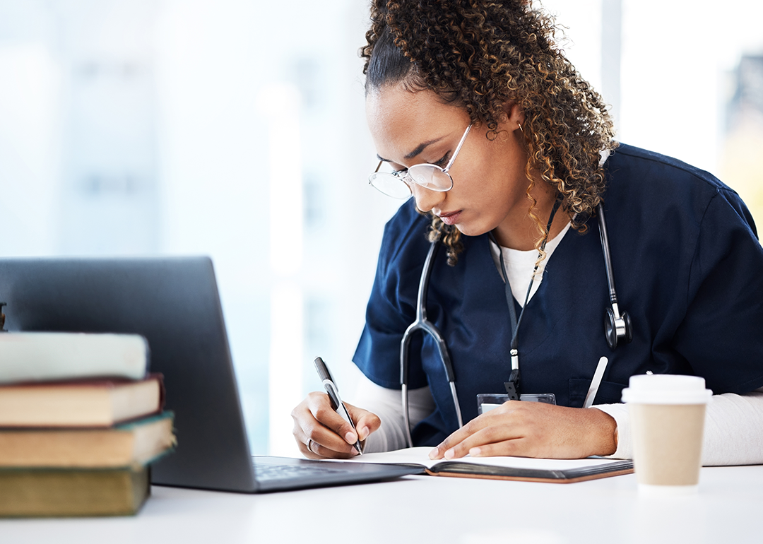 Medical student working at desk, writing in a notebook in front of a laptop, books, and a to-go coffee cup. 