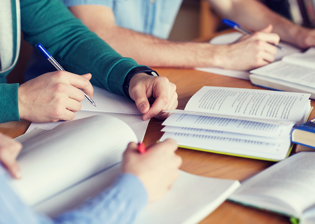 A close up of students arms and hands writing in notebooks and reading textbooks open on a table.