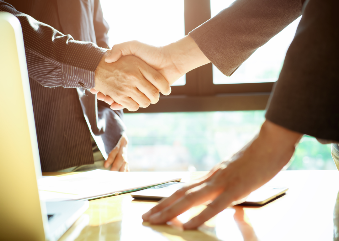 A handshake takes place over a desk and a laptop.
