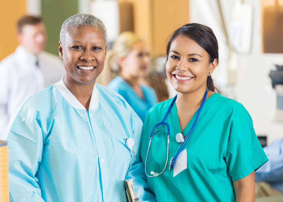 A Black and a Latina nurse in scrubs stand side by side smiling
