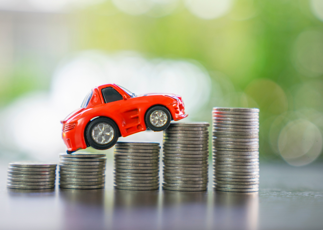 A red toy car rests on stacks of coins that steadily grow taller, signifying a rise in costs.
