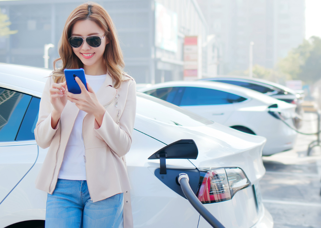 A woman looks at her smartphone while her white electric car charges at a public charge port.