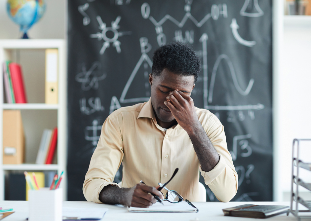 A young teacher looking concerned sitting at the desk in a classroom.