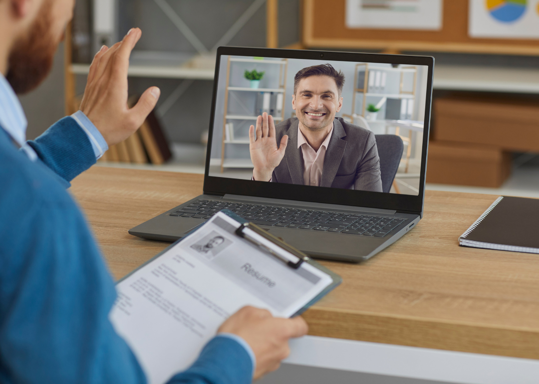 A office worker conducts a video job interview with a man shown on a laptop screen, while holding a copy of the man's resume