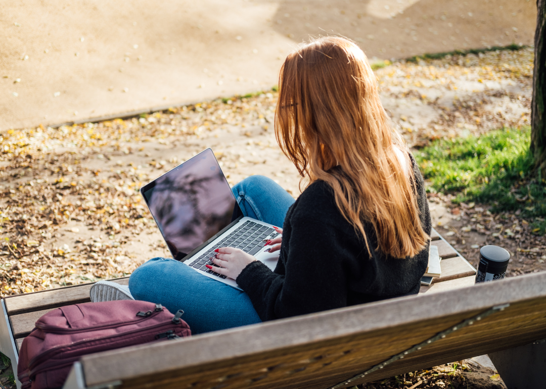 A young woman sits outside on a bench working on a laptop