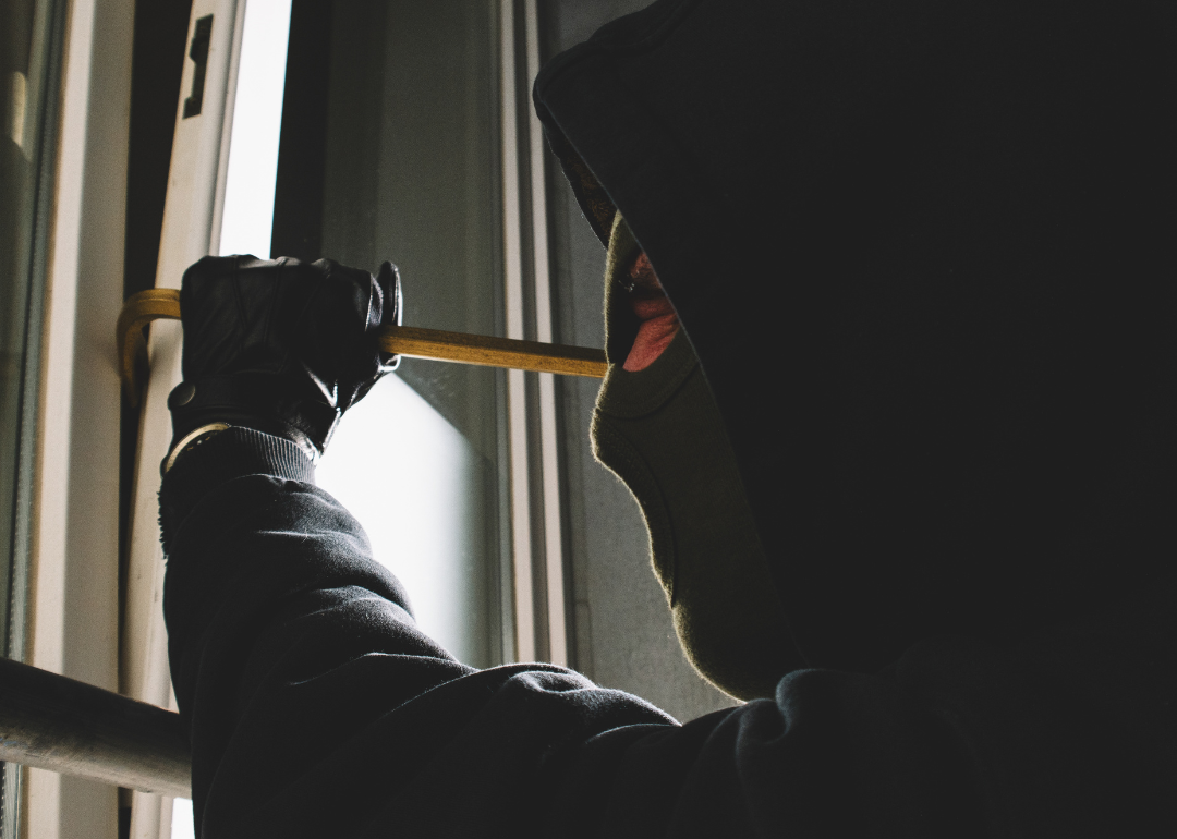 A burglar clad in black uses a crowbar to break into a home through a window casing