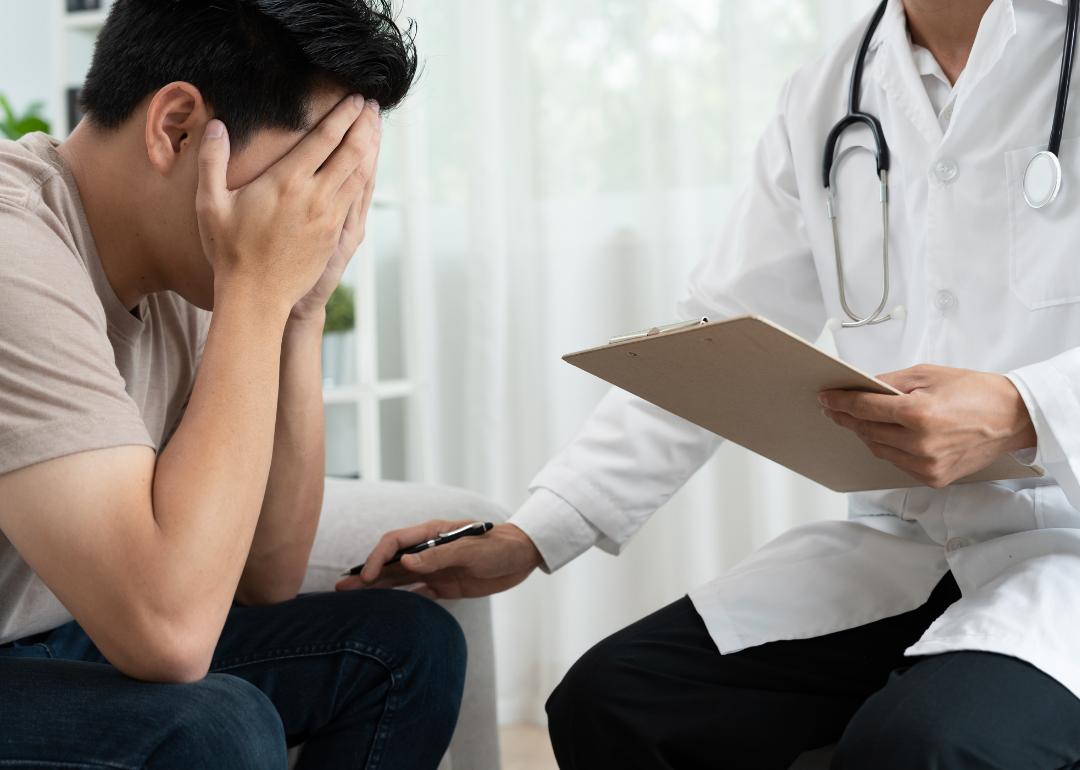 A man sits with his head in his hands, being consulted by a psychologist