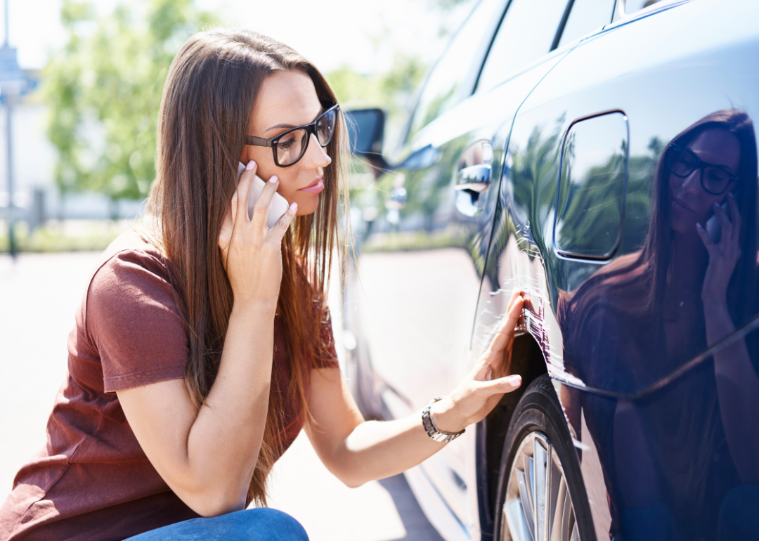 A young woman with glasses inspects damage to the side of her car and makes a phone call